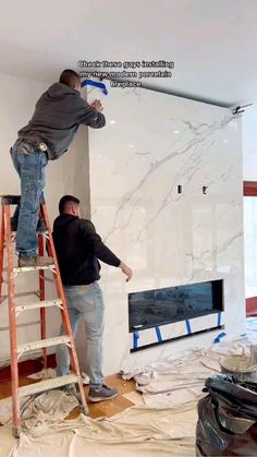 two men working on a wall in a room with white walls and wood floors, while another man stands on a ladder