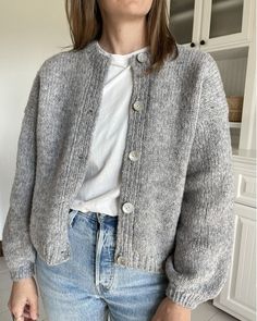 a woman standing in a kitchen wearing a gray cardigan sweater and blue denim jeans