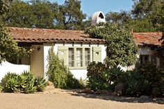 a white house with green shutters next to trees and bushes in front of it