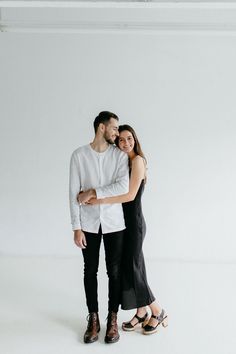 a man and woman standing next to each other with their arms around each other in front of a white wall