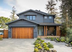 a large gray house with wood garage doors on the front and side of it, surrounded by greenery