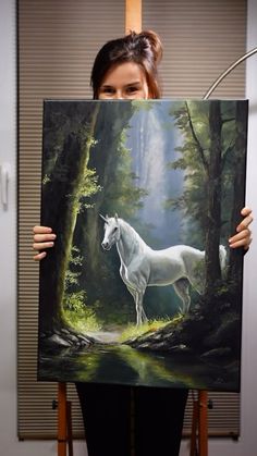 a woman holding up a painting with a white horse in the forest on it's side