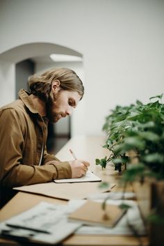 a man sitting at a desk writing on a piece of paper next to a potted plant