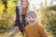 a little boy laughing while standing next to a woman in the woods with trees behind him