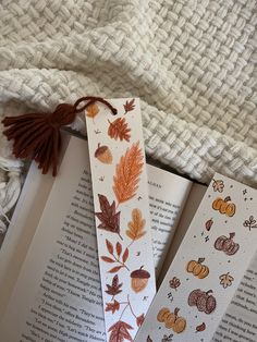 two bookmarks with autumn leaves and acorns are laying on top of an open book