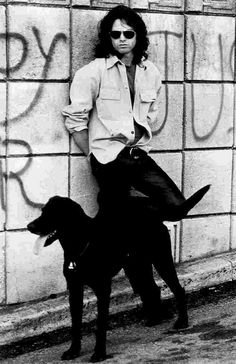 a man standing next to a black dog in front of a wall with graffiti on it