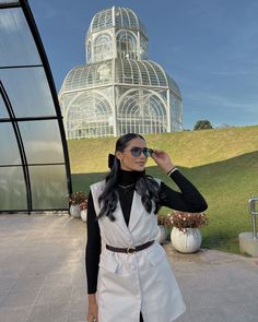 a woman standing in front of a glass house wearing a white vest and black turtle neck sweater