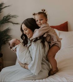 a woman holding a baby on top of her back while sitting on a bed next to a potted plant