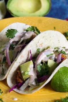 two tacos on a yellow plate with avocado and cilantro in the background