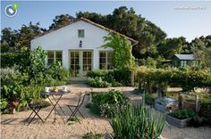 a small white house surrounded by trees and plants in front of a graveled area