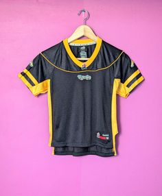 This youth jersey features the Hamilton Tiger Cats (Ticats) of the Canadian Football League. This jersey was made by Reebok in the late-2000s and features a black body with yellow stripes on the sleeves and down each side with Ticat logo patches. This jersey is in excellent condition with no marks, stains, or major condition issues. Size: Youth M/M 15.5" wide from armpit to armpit 21.5" long from top of the back of the collar to bottom (through centre) Sporty Football Season Sports Jersey, Sporty Jersey For Football Season, Sporty Jersey With Team Logo, Sportswear Jersey For Sports Season, Sporty Fitted T-shirt For Football Season, Black Throwback Sports Top, Throwback Black Sports Top, Sporty Team Logo Activewear For Sports, Casual Training Jersey For Sports Season