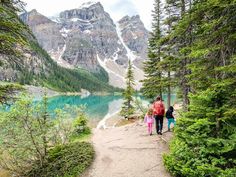 two people and a child are walking on a trail near a lake with mountains in the background