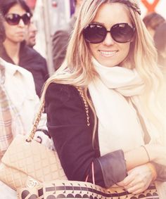 a woman in sunglasses carrying a large handbag and smiling at the camera with other people behind her