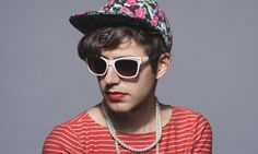 a young man wearing sunglasses and a hat with flowers on it, sitting in front of a gray background