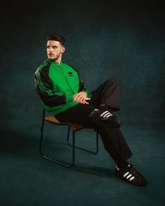 a man sitting in a chair wearing a green jacket and black sweatpants with his legs crossed