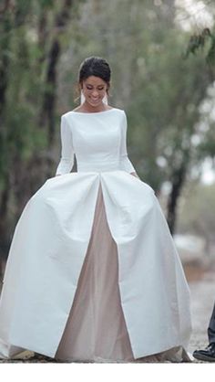 a woman in a white wedding dress walking down a dirt road with trees behind her