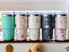 a teddy bear sitting next to a row of travel mugs with designs on them