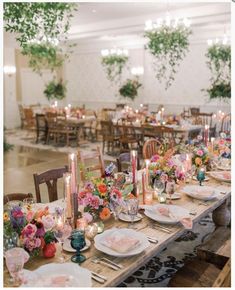 a long table with plates and candles on it in a room filled with flowers, greenery and hanging plants