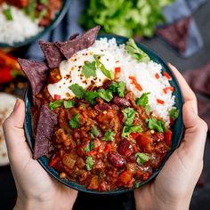 two hands holding a bowl of chili and rice with tortilla chips on the side