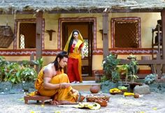 a man sitting on the ground in front of a house next to a woman standing behind him