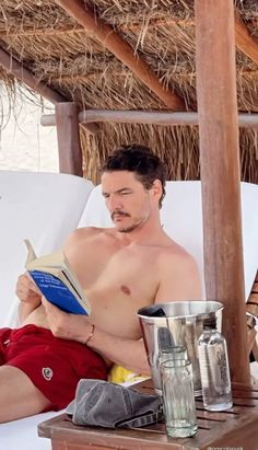 a shirtless man reading a book while sitting in a chair under a straw hut