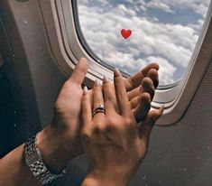 two hands reaching out an airplane window to touch the sky with a red heart on it