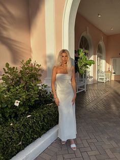 a woman in a white dress is standing on a brick walkway near bushes and trees