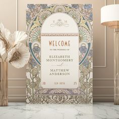 an ornate welcome card on a marble table next to a lamp and vase with flowers