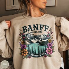 Celebrate one of the most pristine places in the Canadian Rocky Mountains, Banff National Park, with our Women's Banff National Park Crewneck from Triangle Threads! This vintage style sweatshirt is perfect to remember that once in a lifetime trip to Banff or to celebrate an upcoming journey! Comfy, soft, and sporting a vintage design with colorful wildflowers and graphic illustration of Lake Moraine itself, this sweatshirt will bring you back to the mountains and lakes of Banff in style! * Produ Spring Outdoor Sweatshirt With Letter Print, Outdoor Letter Print Sweatshirt For Spring, National Park Shirts, Lake Moraine, National Park Sweatshirt, Colorful Wildflowers, Canadian Gifts, Wildflower Shirt, To The Mountains