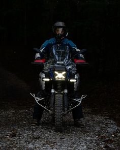 a man riding on the back of a motorcycle in the woods at night with its lights on