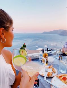 a woman sitting at an outdoor table with food and drinks in front of the ocean