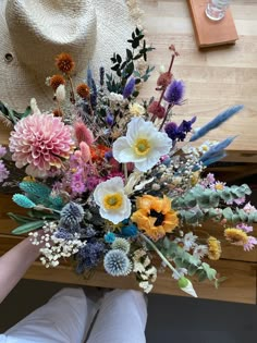 a person holding a bouquet of flowers on top of a wooden table next to a straw hat