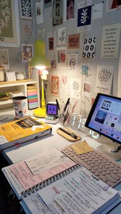 a desk with a computer, notebooks and other items on it in front of a wall full of pictures