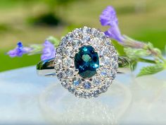 a blue diamond ring sitting on top of a table next to purple flowers and green leaves