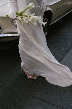 a woman in a white dress is walking by a car with flowers on her arm