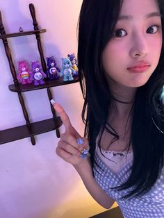 a woman with long black hair pointing at the camera while standing in front of a shelf filled with small teddy bears