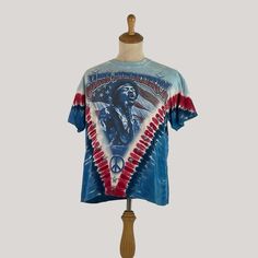 Step into the music scene with this Vintage Jimi Hendrix American Flag Blues Guitarist Tie Dye Tee. Officially licensed and medium-sized, this shirt is a genuine tribute to the legendary guitarist. Its tie-dye pattern with the American flag motif channels the hippie vibes of the era. Ideal for music enthusiasts, collectors, and those who appreciate vintage fashion, this tee is more than apparel; it's a piece of concert memorabilia representing a crucial moment in music history. Whether you're a Band Merch Blue T-shirt With Sublimation Print, Blue Screen Print T-shirt For Concert, Hippie Short Sleeve T-shirt For Music Festival, Blue Band Merch T-shirt, Blue Band Merch Top With Sublimation Print, Blue Graphic Print T-shirt For Festivals, Blue Cotton T-shirt For Festivals, Retro Short Sleeve T-shirt For Festival, Festival Fan Merchandise Short Sleeve Tops