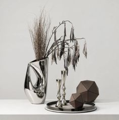 two silver vases sitting on top of a white table next to each other with dried plants in them