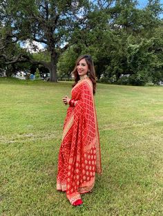 a woman in a red and gold sari standing on the grass with trees behind her