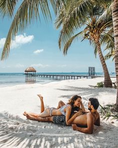 a man and woman laying on the sand under palm trees in front of an ocean pier