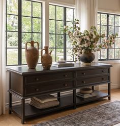 two vases are sitting on a table in front of large windows with white curtains