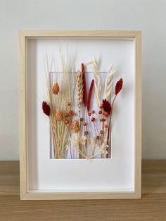 an image of dried flowers in a white frame on a wooden table with a wall behind it