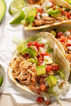 three tacos with shredded meat, tomatoes and avocado on paper wrappers