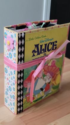 an open alice book sitting on top of a wooden table next to a pink ribbon