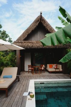 an outdoor deck with lounge chairs and umbrellas next to a swimming pool in front of a thatched roof hut