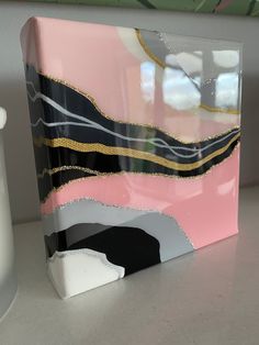 a pink, black and gold painting on a white surface next to a cup with a vase