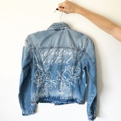 a denim jacket with flowers on it hanging from a hook and someone's hand holding the hanger