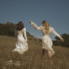 two women in white dresses are walking through tall grass and one is pointing to the sky