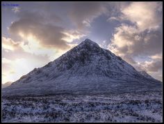 a snow covered mountain with clouds in the sky above it and some grass on the ground