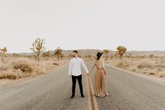 a man and woman holding hands while walking down the middle of an empty desert road